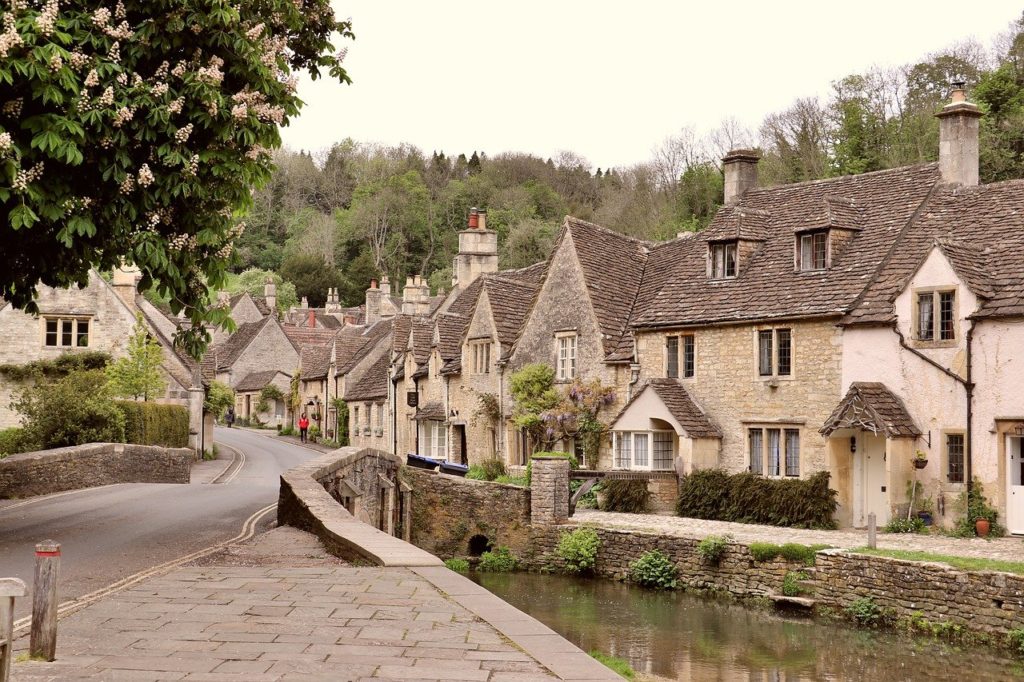image of an English street with stone brick houses, a small river running thorugh the middle and green trees in the background.