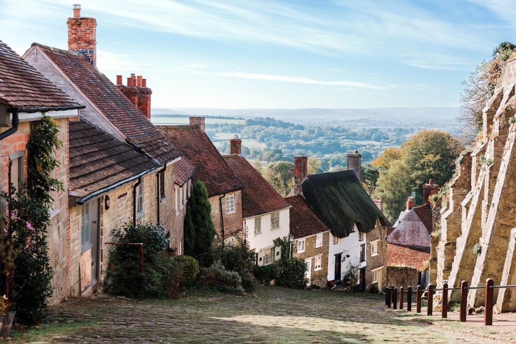 Image looking down a peaceful English street with cobbled streets, brick houses and green hills in the distance with a blue sky backdrop
